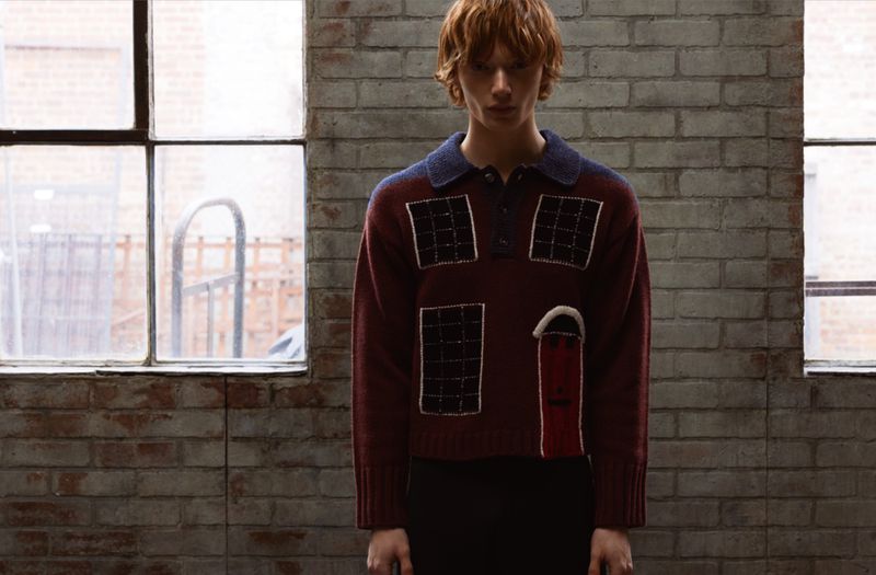 Male fashion model poses indoors wearing a burgundy sweater with a blue collar against a brick wall and window, medium shot.
