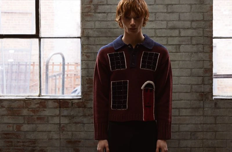 Male fashion model poses indoors wearing a burgundy sweater with a blue collar against a brick wall and window, medium shot.