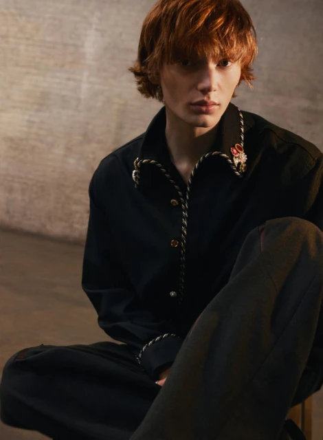 Person with red hair sitting indoors wearing a black shirt with decorative trim. Neutral wall backdrop with soft lighting.