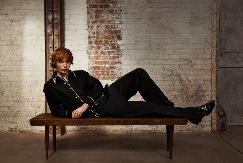 An adult man with auburn hair, dressed in a black outfit, is lounging on a wooden bench indoors in front of a distressed brick wall.