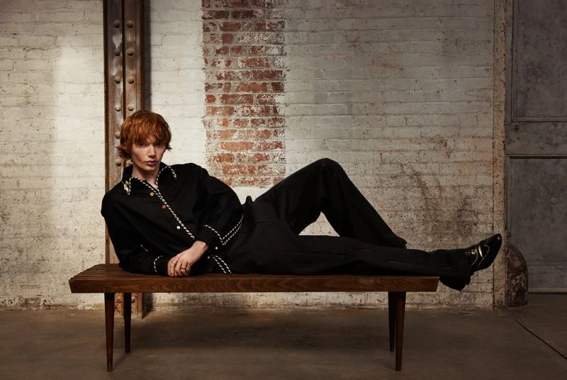 An adult man with auburn hair, dressed in a black outfit, is lounging on a wooden bench indoors in front of a distressed brick wall.