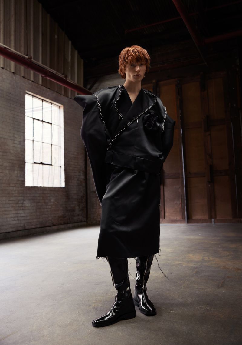 Fashion forward young adult in an all-black, oversized outfit and patent leather boots, posing in a raw, industrial-style warehouse space.