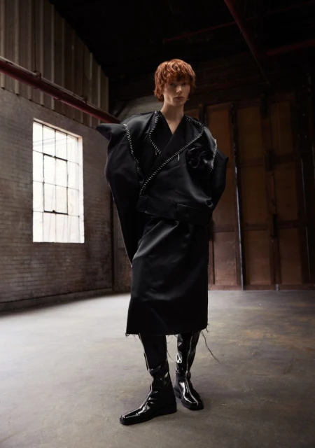 Fashion forward young adult in an all-black, oversized outfit and patent leather boots, posing in a raw, industrial-style warehouse space.