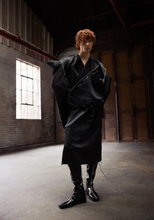 Fashion forward young adult in an all-black, oversized outfit and patent leather boots, posing in a raw, industrial-style warehouse space.