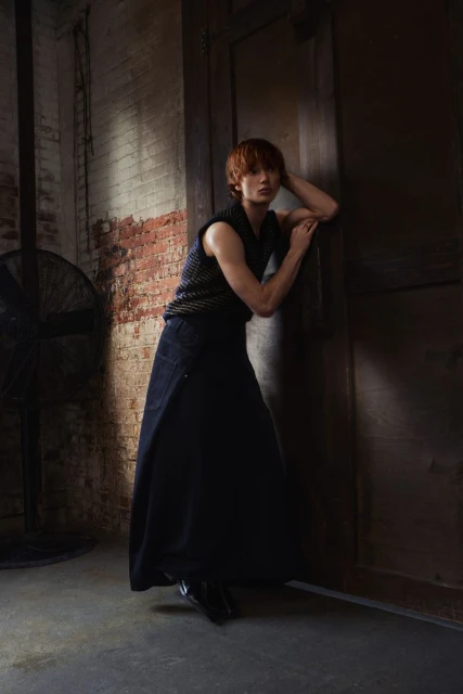 Woman in a sleeveless top and long skirt leaning against a doorway in a dimly lit interior with brick and concrete walls.