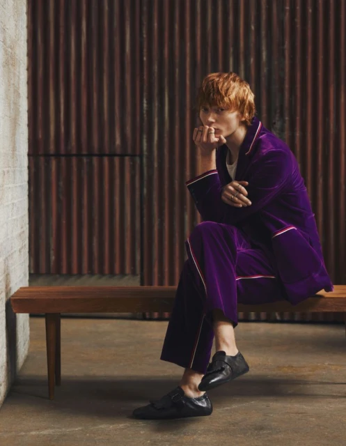 Adult male with reddish hair sitting on wooden bench in purple velvet suit with black dress shoes in indoor setting with brick wall.