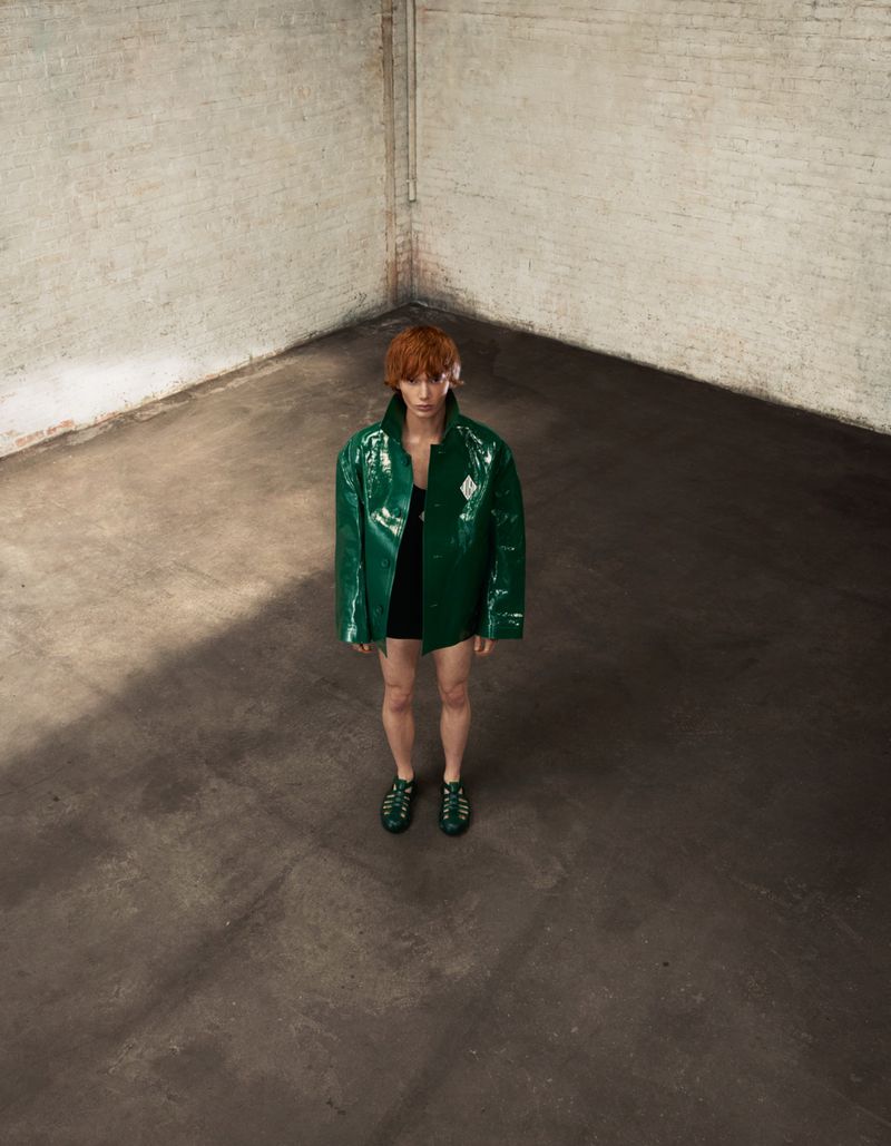 A person with auburn hair stands in a grungy studio wearing an oversized, shiny green vinyl jacket and matching gladiator sandals.
