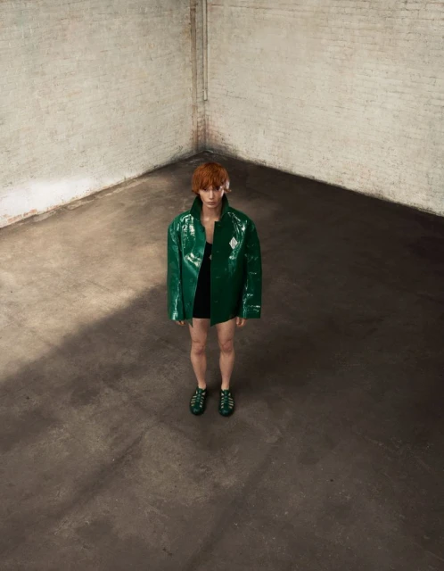 A person with auburn hair stands in a grungy studio wearing an oversized, shiny green vinyl jacket and matching gladiator sandals.