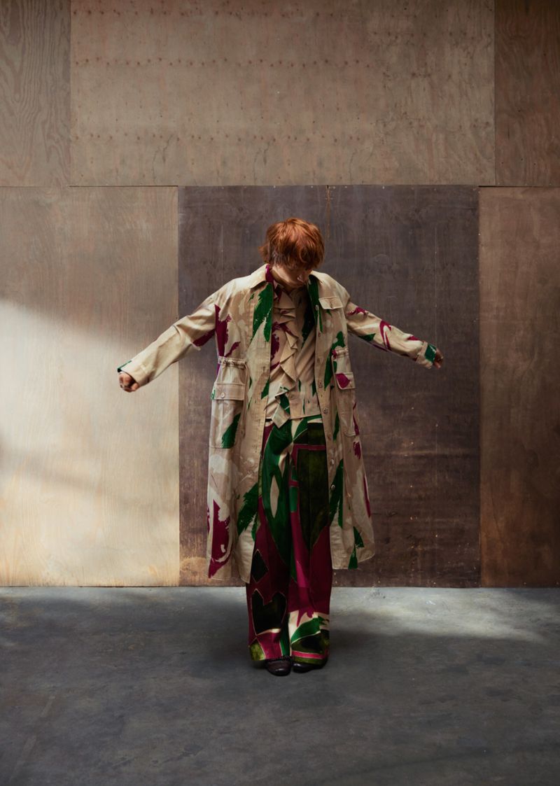 Fashionable person with red hair in printed coat and trousers stands in muted room with concrete floor and neutral color-blocked walls.