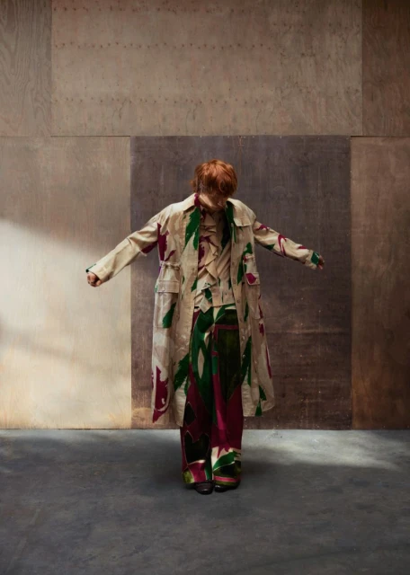Fashionable person with red hair in printed coat and trousers stands in muted room with concrete floor and neutral color-blocked walls.
