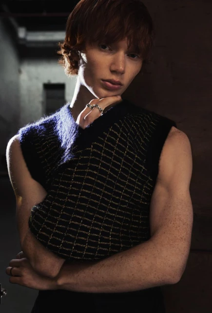 Studio portrait of a young redhead man wearing a black vest with arms crossed, looking at the camera against a dark background.