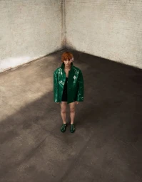 Model wearing a glossy green jacket standing in an empty industrial warehouse with exposed brick walls.