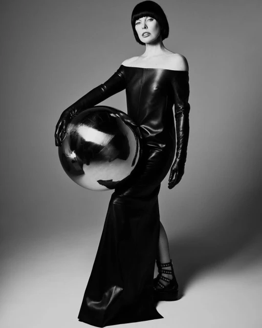 Monochrome portrait of a woman in a black off-the-shoulder dress and long gloves, holding a large reflective sphere in a studio.