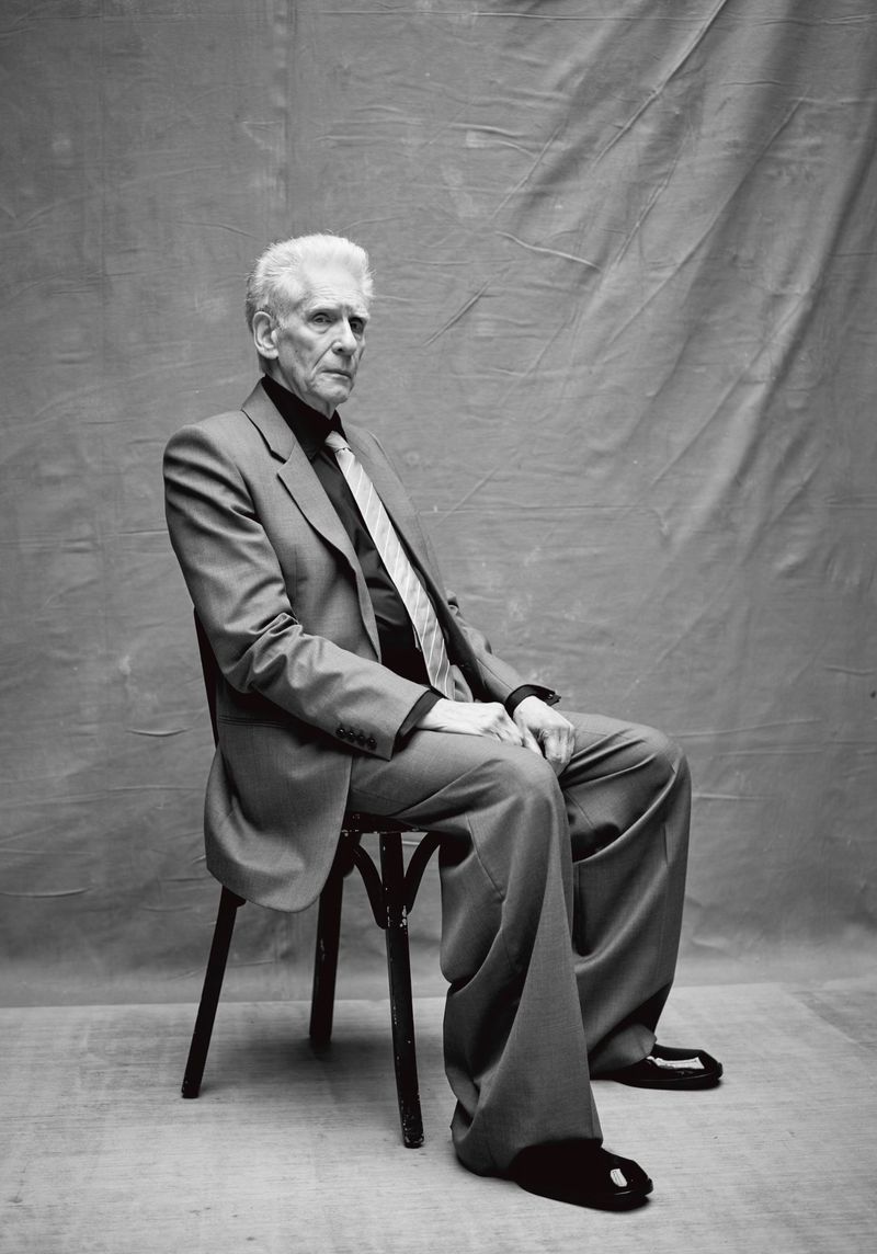 Elderly man in a suit sitting on a chair against a draped backdrop. Black and white studio portrait, full shot, professional attire.