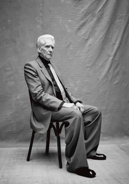 Elderly man in a suit sitting on a chair against a draped backdrop. Black and white studio portrait, full shot, professional attire.