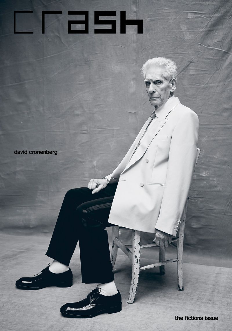 Elderly man seated in front of gray backdrop for magazine cover. He wears a white blazer, dress pants, and shiny black dress shoes.