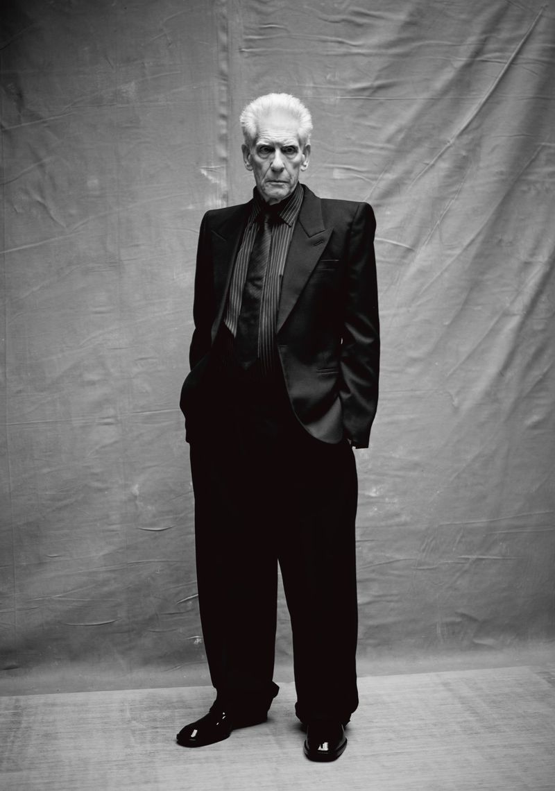 Full-length portrait of an elderly man with white hair in a black suit, striped shirt and tie, standing indoors against a grey backdrop.