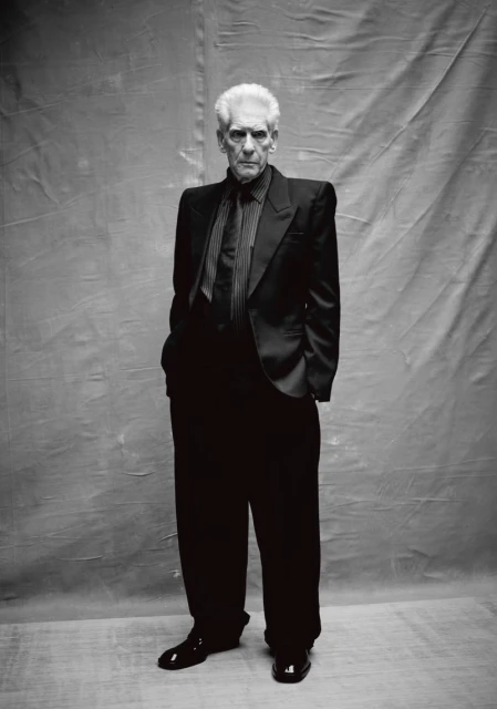 Full-length portrait of an elderly man with white hair in a black suit, striped shirt and tie, standing indoors against a grey backdrop.