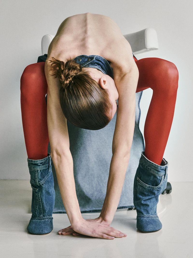 Artistic studio shot of a human figure arching backward wearing denim boots, red tights, and a denim skirt against a light backdrop.