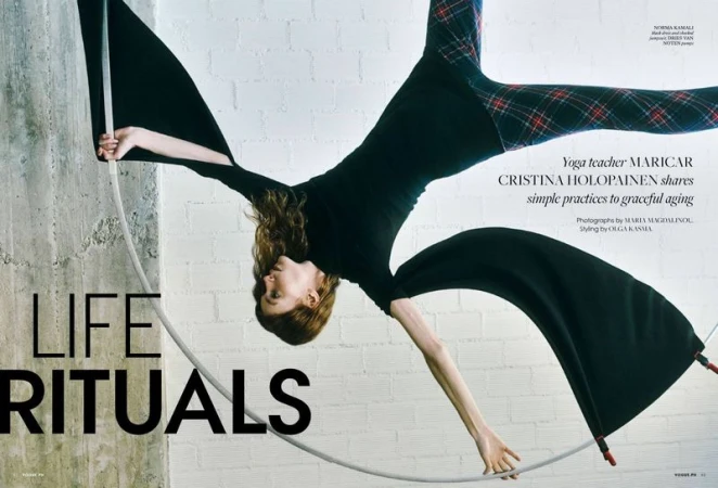 A woman performs aerial yoga while suspended upside down in a hoop wearing a long-sleeved black shirt and plaid pants against concrete.