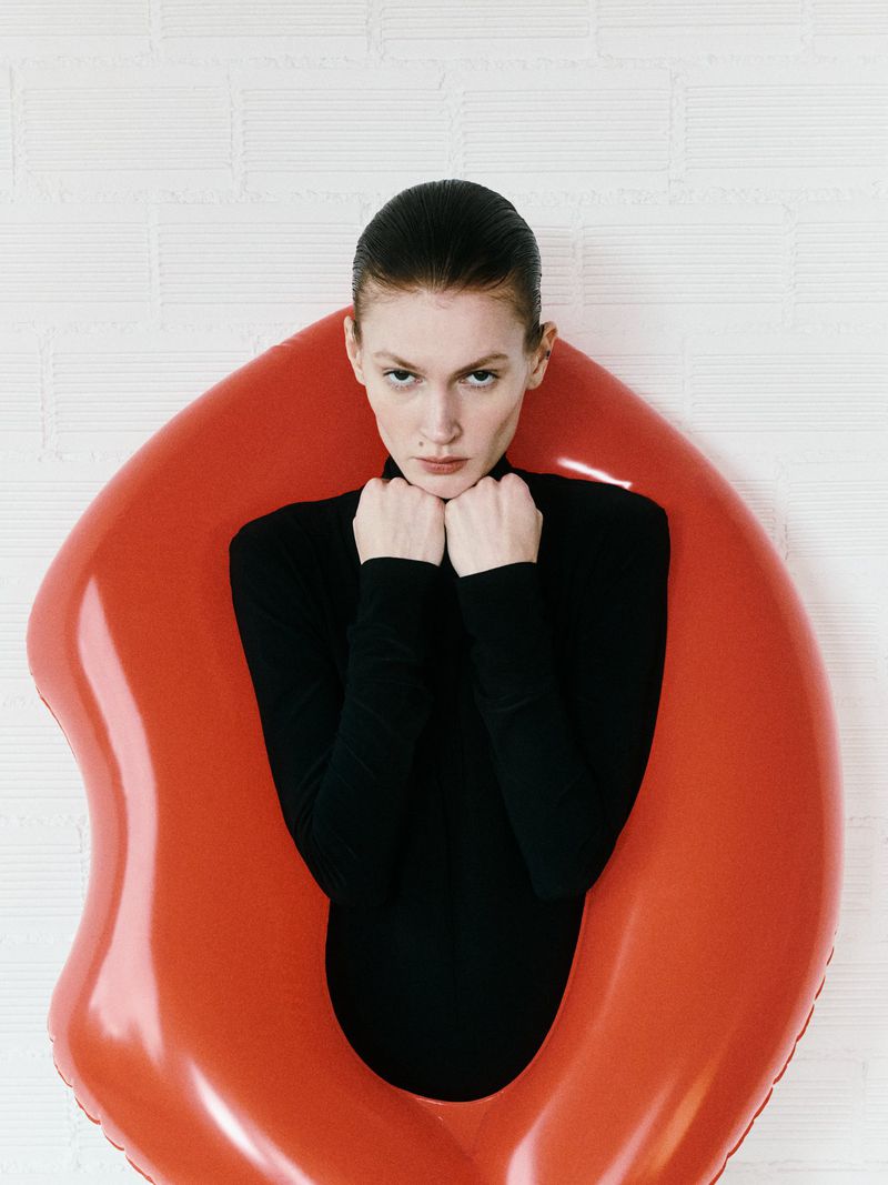 Woman in a black turtleneck seated within a bright red, inflatable chair against a white, patterned backdrop.