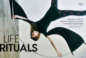 Woman hanging suspended upside down holding a curved metal ring against a white brick wall and concrete pillar background.