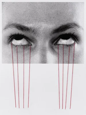 Close-up black and white portrait of a person's eyes with red thread sewn down from the lower eyelids onto a plain paper background.