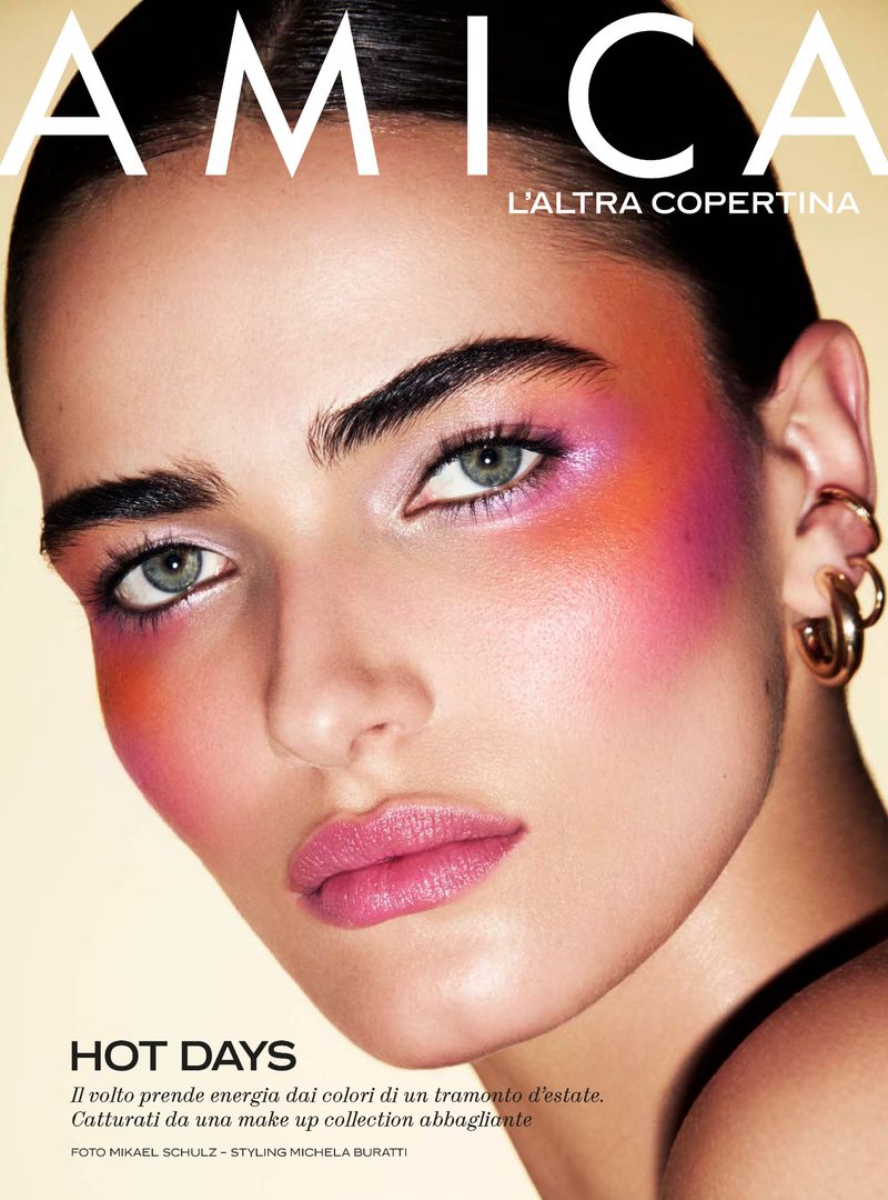 Close-up of a young adult woman with bold pink and orange makeup, gold hoop earrings, and dark hair, set against a neutral background.