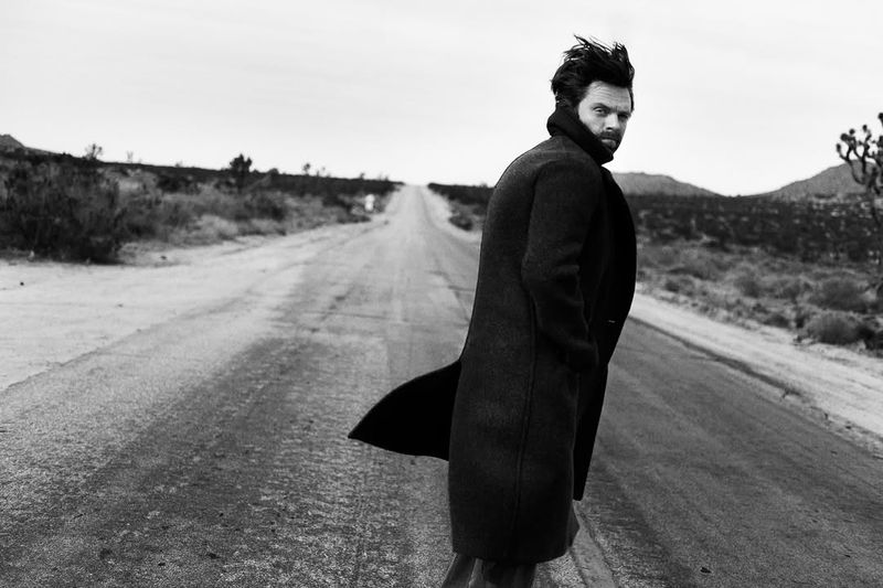 Monochrome portrait of a man with dark hair and a beard, wearing a coat, looking over his shoulder on an empty desert road.