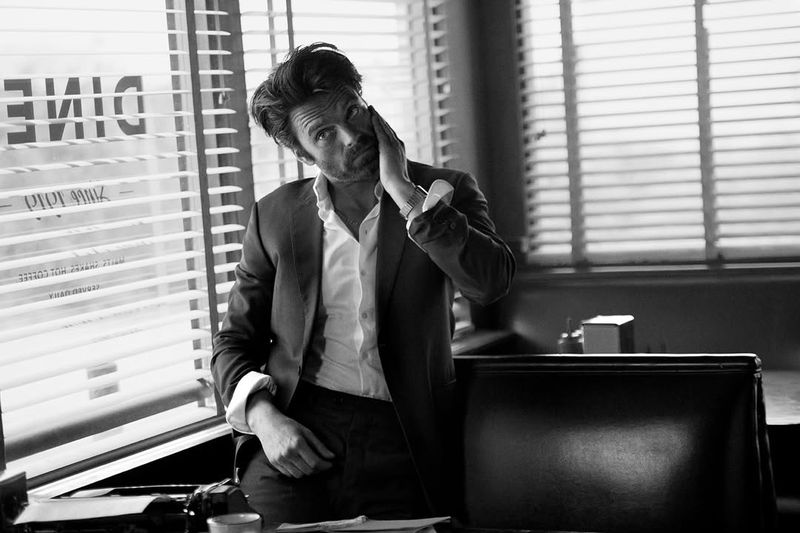 Actor Sebastian Stan in a dark suit jacket and white shirt leaning against a booth in a diner
