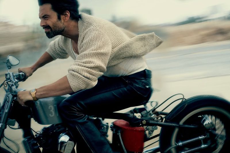 Adult male rides a vintage motorcycle on an open road; he wears a beige sweater and dark pants in a daytime outdoor scene.