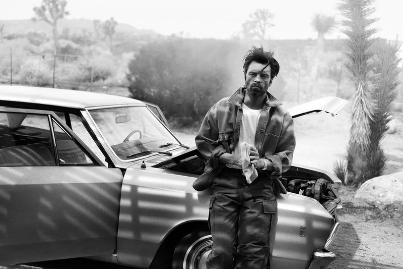 Monochrome shot of a man with a cloth leaning on a vintage car with the hood up, set in a desert landscape with scrub vegetation.