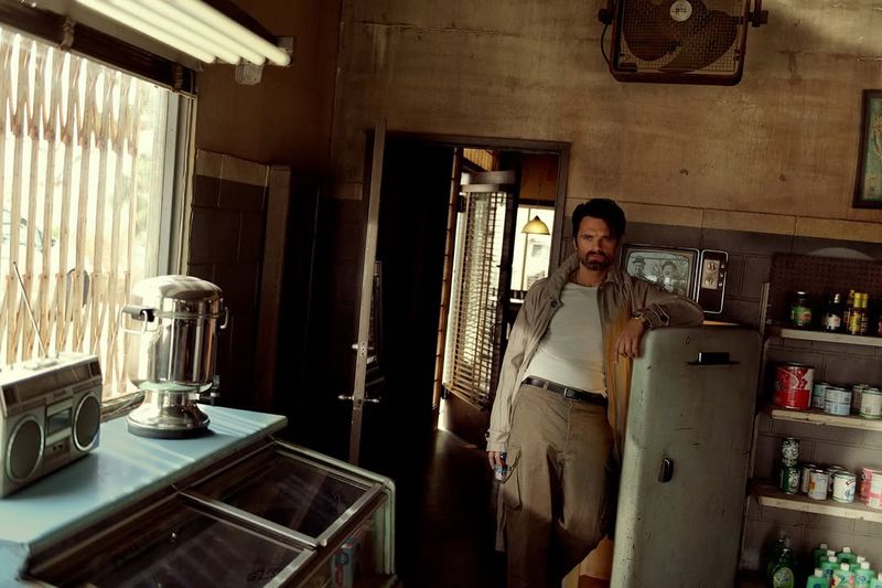 A man in a trench coat leans against a refrigerator in a vintage-styled diner with shelves of goods, a boombox, and dim, atmospheric lighting.