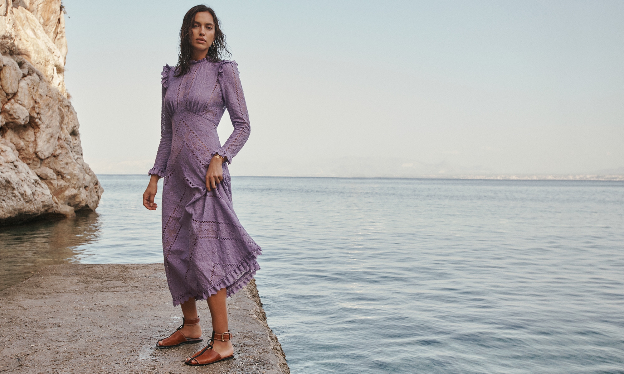 Model Irina Shayk wearing a long purple lace dress standing on a concrete pier by the ocean.