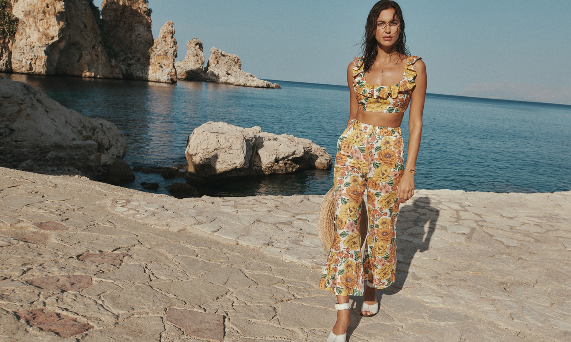 Woman in floral crop top and pants on stone beach with rock formations, wearing white sandals, against a blue sky and ocean backdrop.