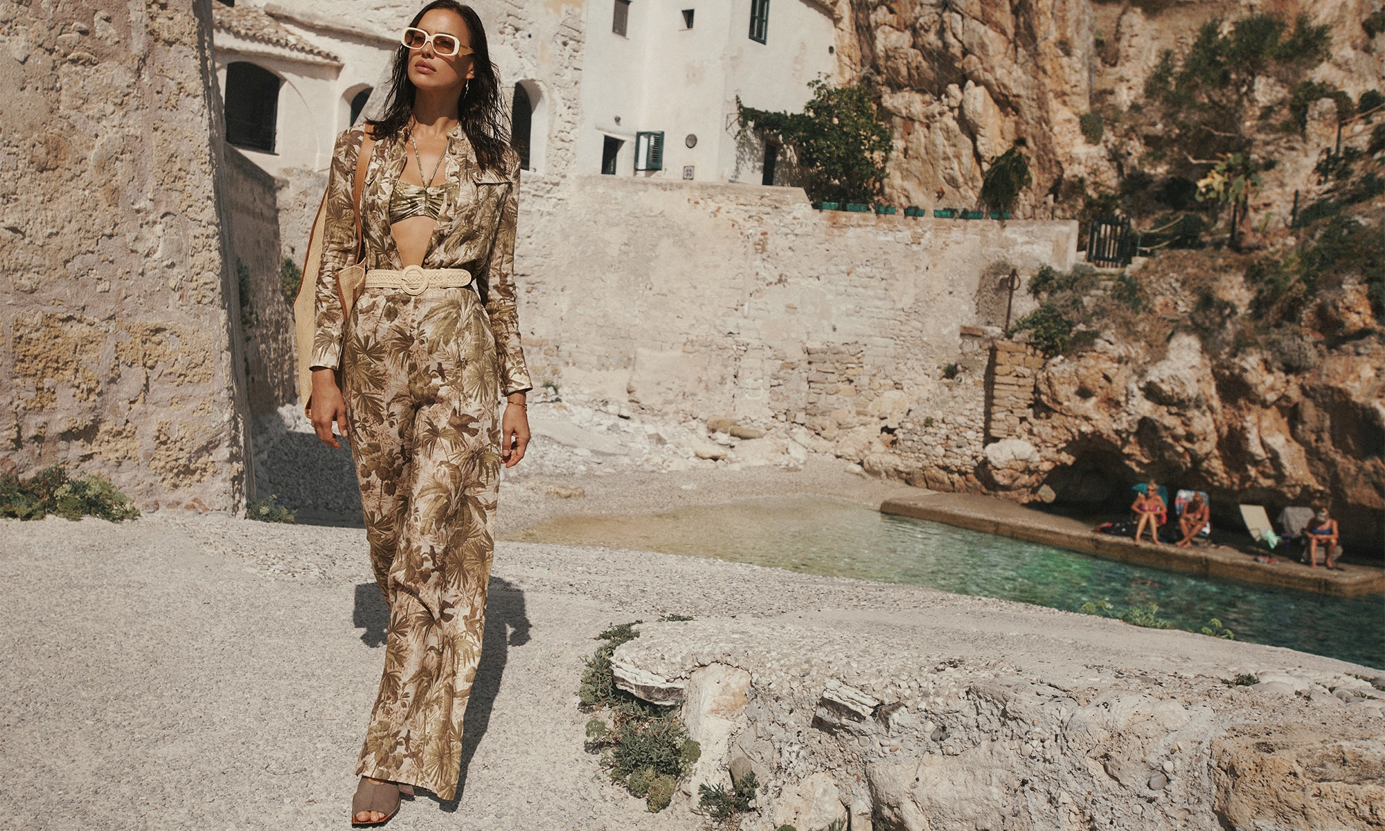 An adult woman in a palm-print outfit walks on a stone path with a bay and rocky cliffs in the background, captured in an eye-level shot.