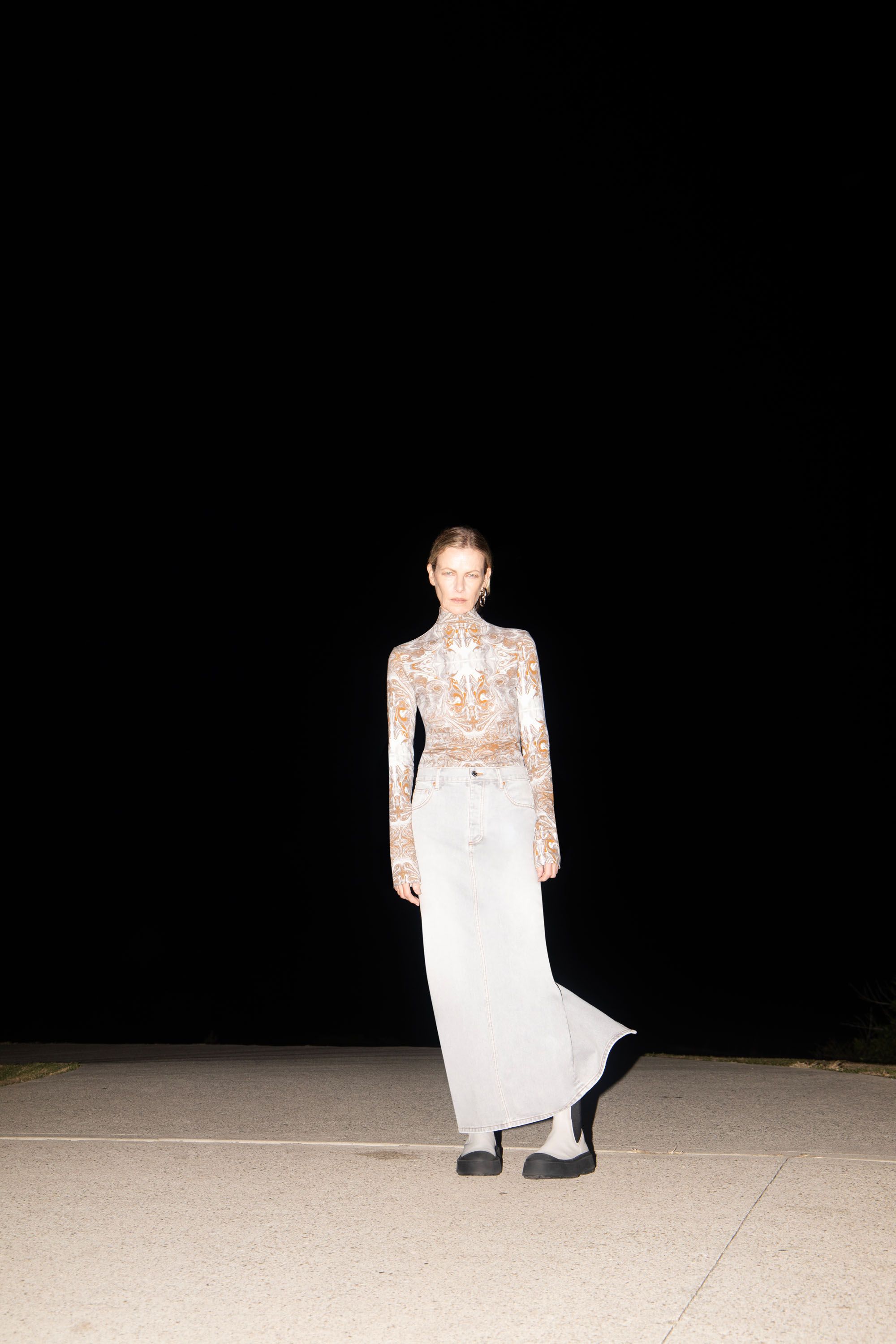 Fashionable woman in patterned top and maxi skirt poses outdoors at night in an editorial style, showcasing modern design and dark mood.