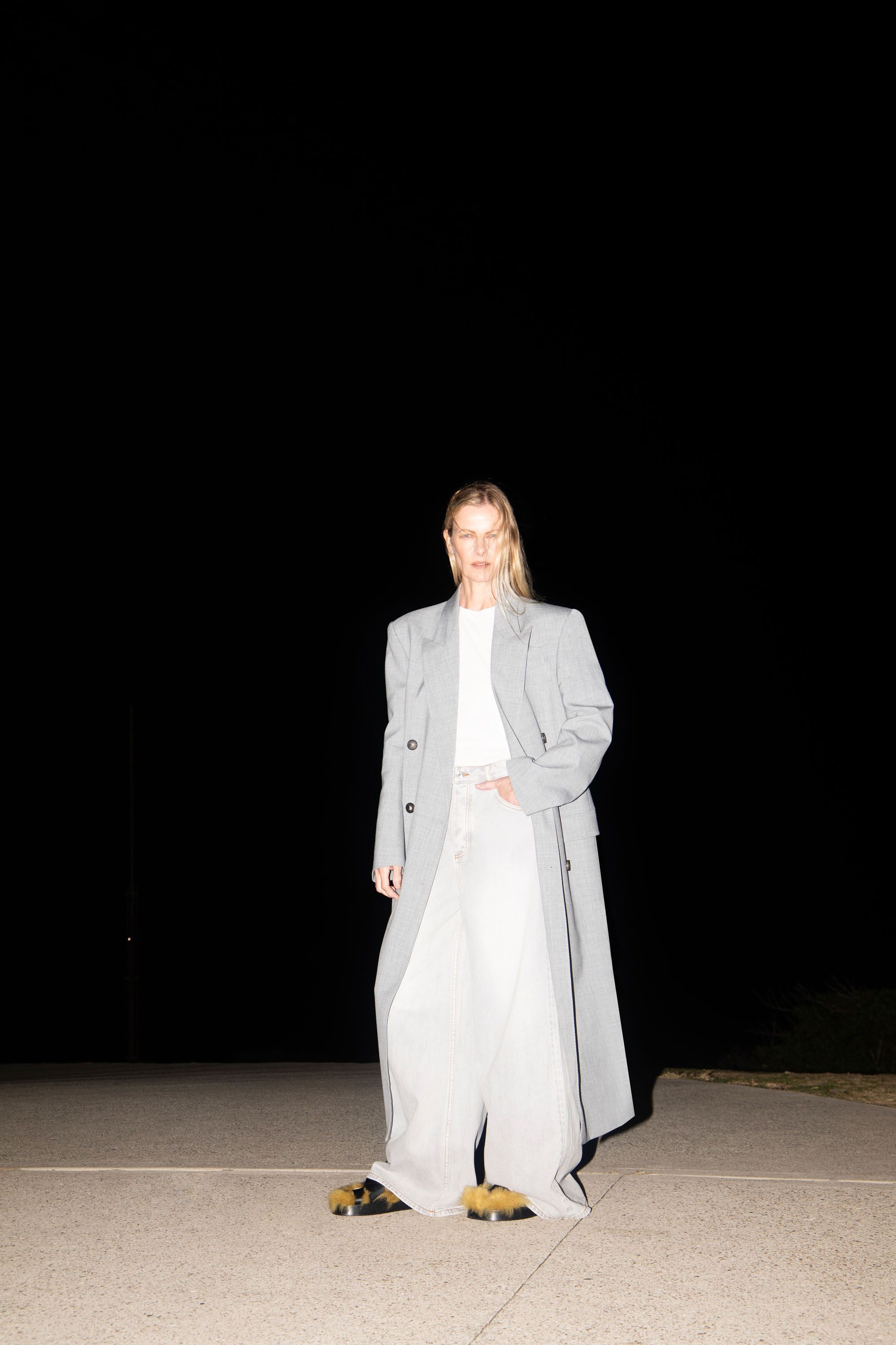 Model standing outdoors at night wearing a grey coat over white top and baggy light trousers.