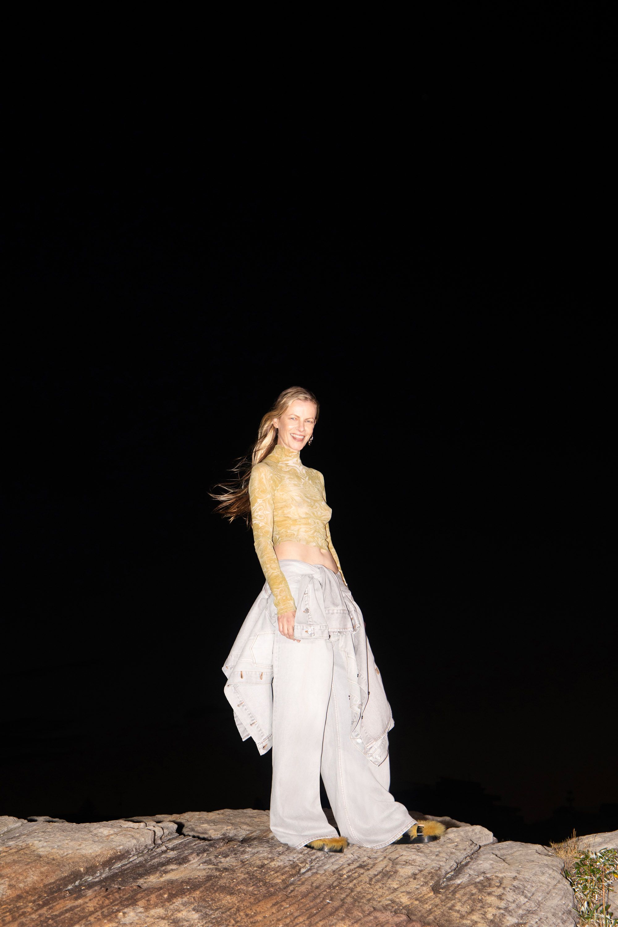 A woman smiles on a rock. She wears a gold top, white pants, and sandals, with a dark background and soft lighting.