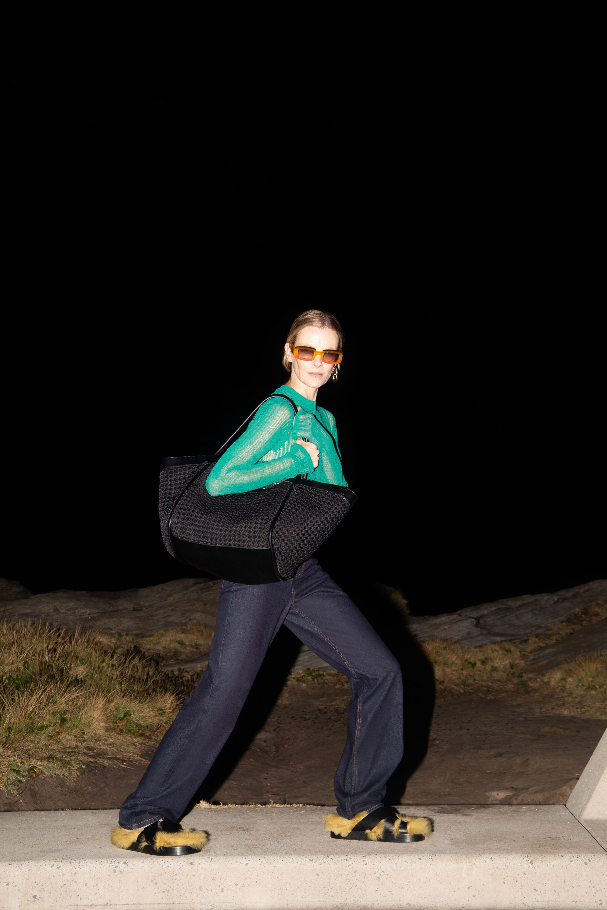 A woman in a green top and sunglasses carrying a black tote bag walks on a concrete path at night, illuminated by artificial light.