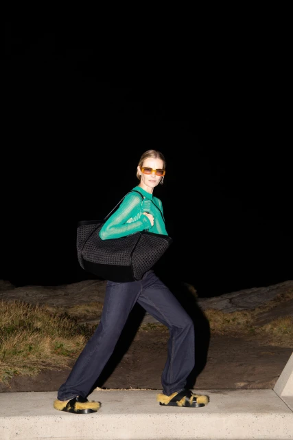 A woman in a green top and sunglasses carrying a black tote bag walks on a concrete path at night, illuminated by artificial light.