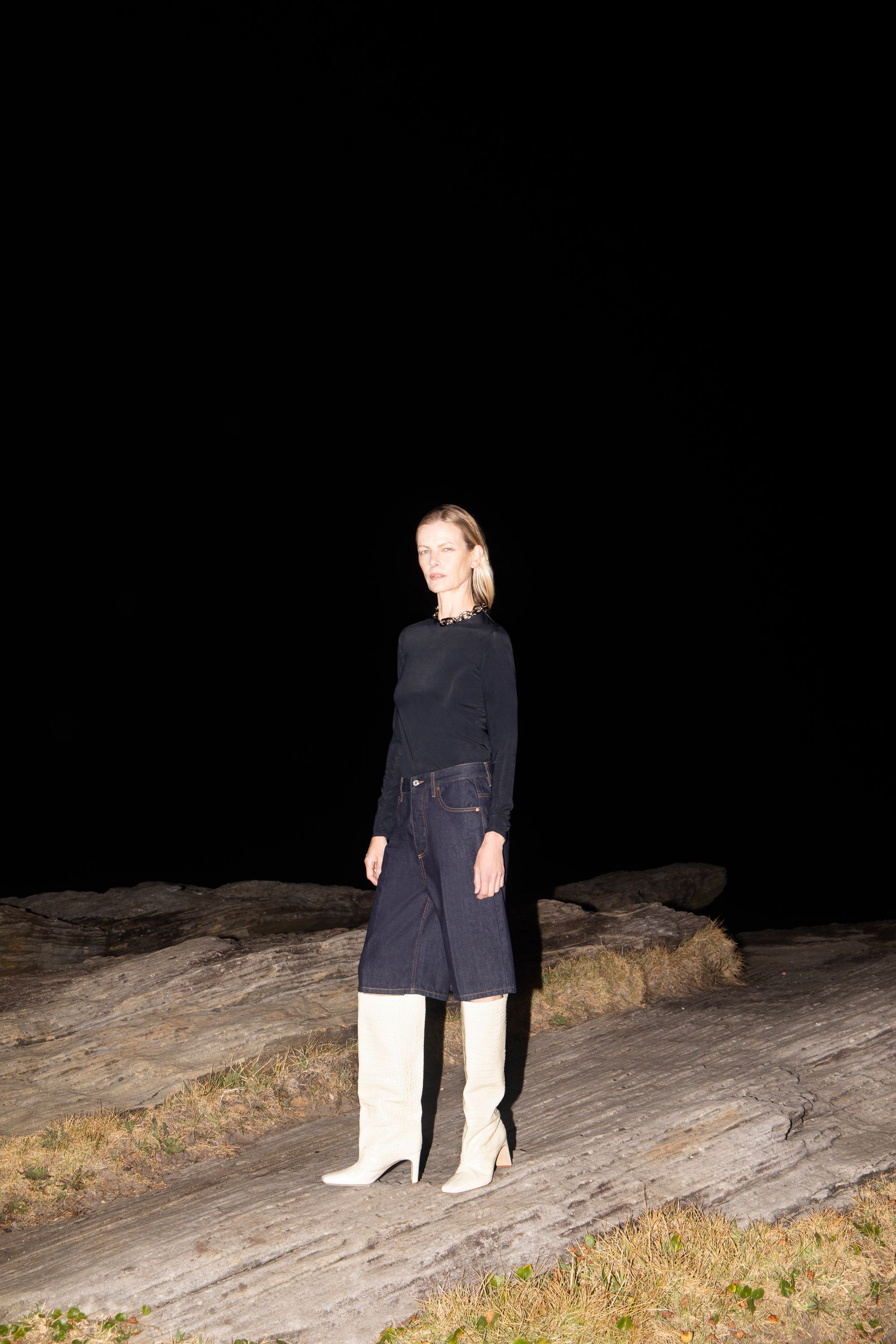 Full-length shot of a blonde adult female in a navy long-sleeve top, dark denim culottes, and tall white boots, standing on a rock formation.