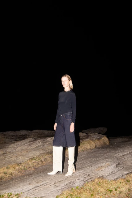 Full-length shot of a blonde adult female in a navy long-sleeve top, dark denim culottes, and tall white boots, standing on a rock formation.