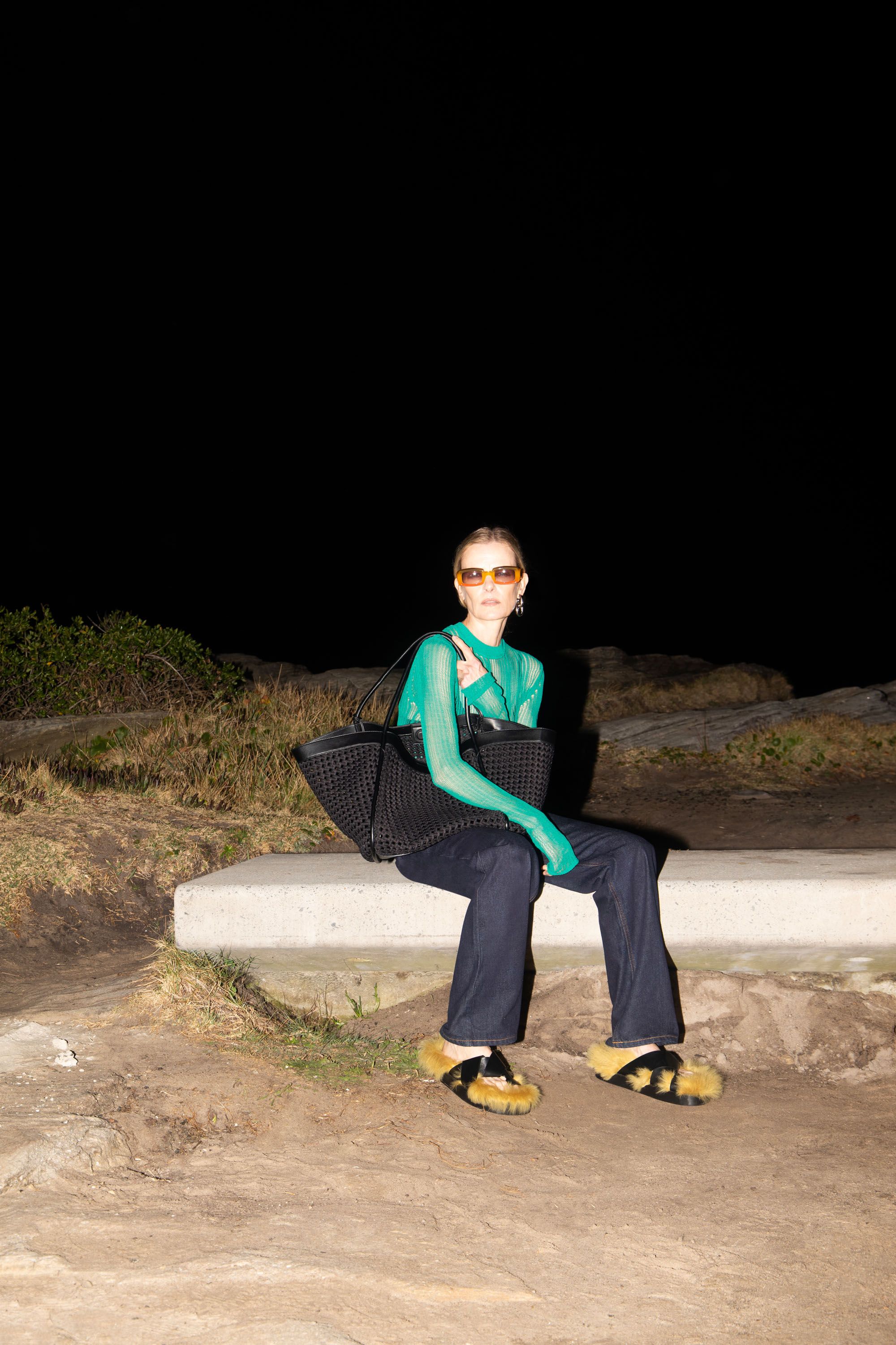 A woman wearing a sheer green top, sunglasses, and slippers sits on a concrete barrier at night, holding a black tote bag.