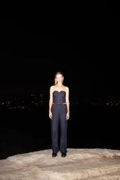 Model standing on a rocky outcrop at night wearing a dark denim strapless top and matching trousers with city lights in the distance