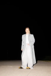 Model standing outdoors at night wearing a grey coat and wide-leg trousers under direct flash lighting.