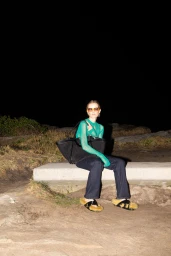 Model seated outdoors at night wearing a sheer green top, dark denim jeans, sunglasses, and holding a large black tote bag