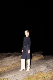 Model standing on rocky coastal terrain at night wearing dark denim trousers and white knee-high boots under direct flash lighting