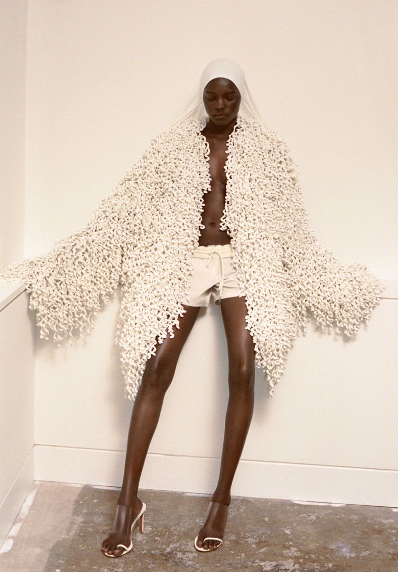 Fashion shot of a Black female model wearing a white headscarf, cream-colored shorts, clear high heels, and a textured fringed coat indoors.