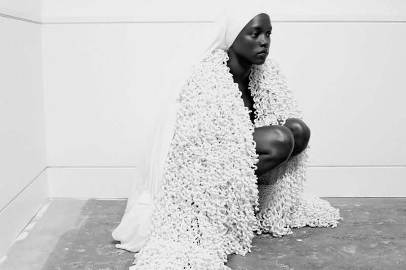 Monochrome portrait of an African woman in a white headscarf, crouching indoors draped in a looped white textile, looking off to the side.